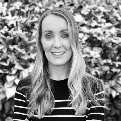 Black-and-white portrait of a person with long wavy hair, wearing a striped top, smiling, with a leafy background.