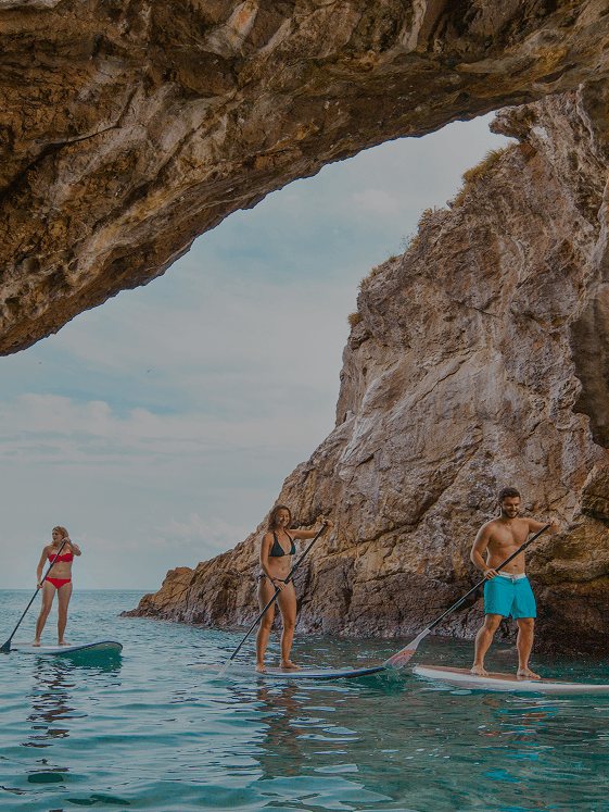 Three people in swimsuits stand-up paddleboarding on turquoise water beneath a large rocky arch.