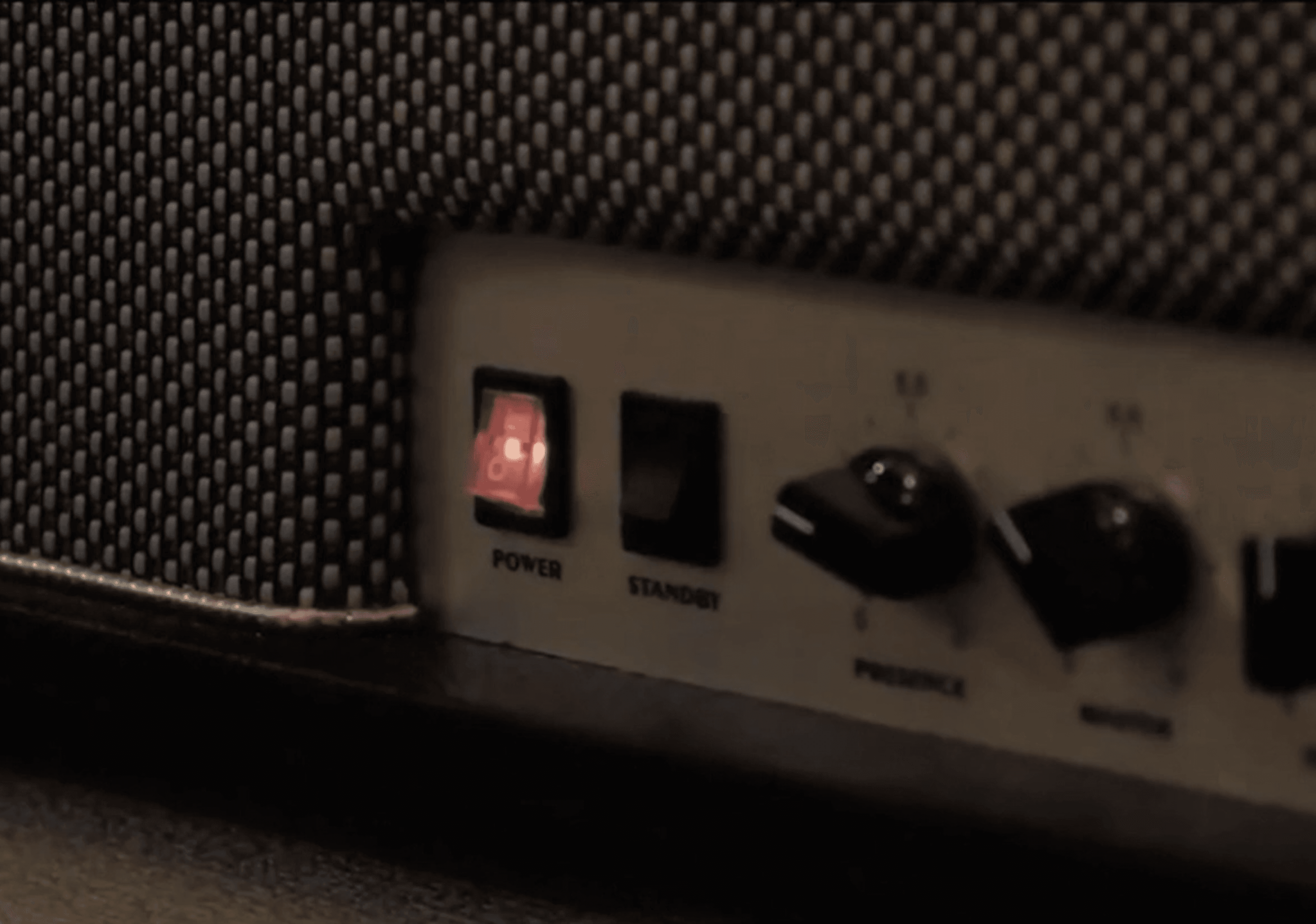 Close-up of a speaker front panel with a glowing red power switch, a standby switch, and two control knobs beside a black mesh grille.
