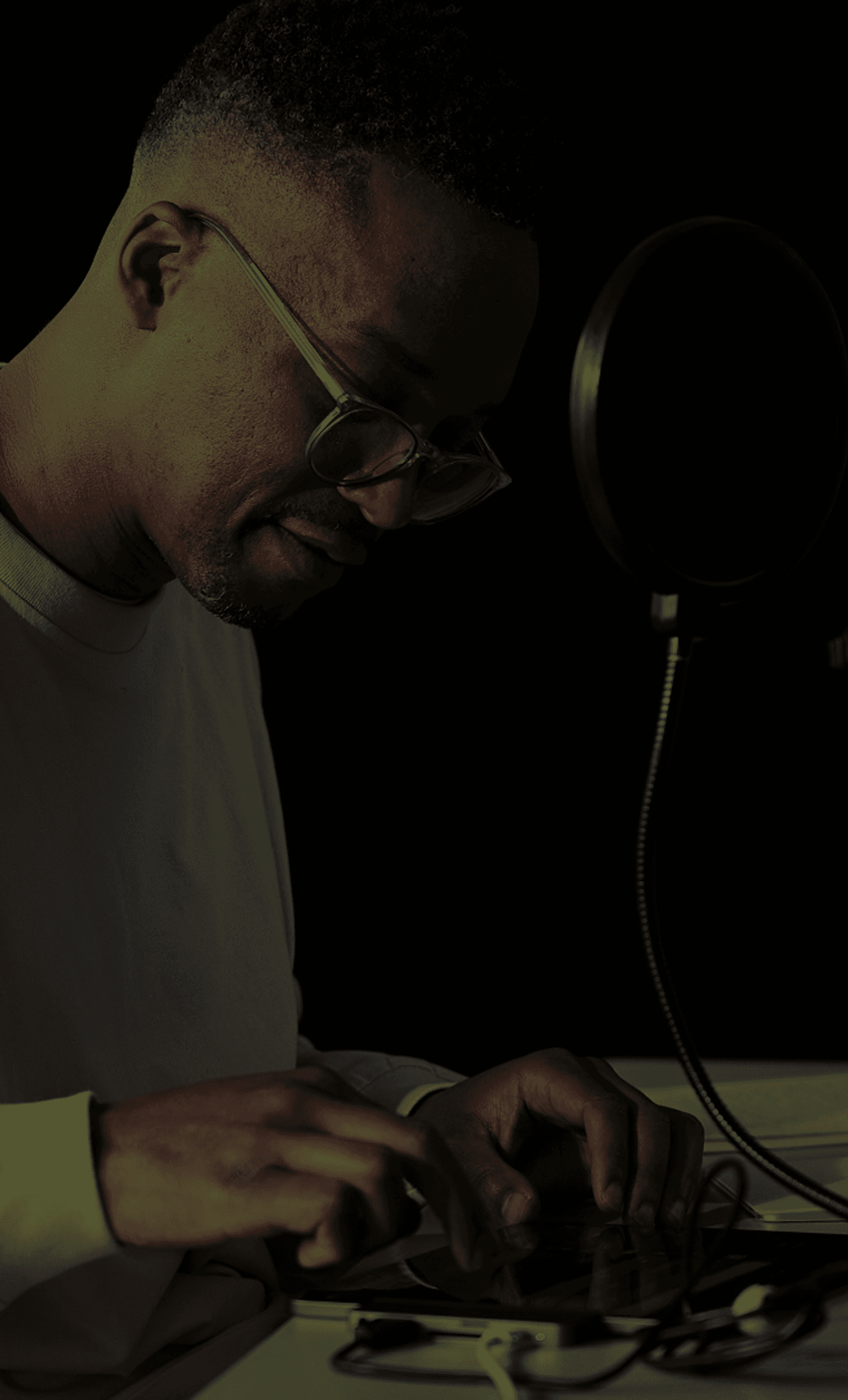 Person wearing glasses sits at a dimly lit desk, typing on a tablet, with a microphone and pop filter nearby.