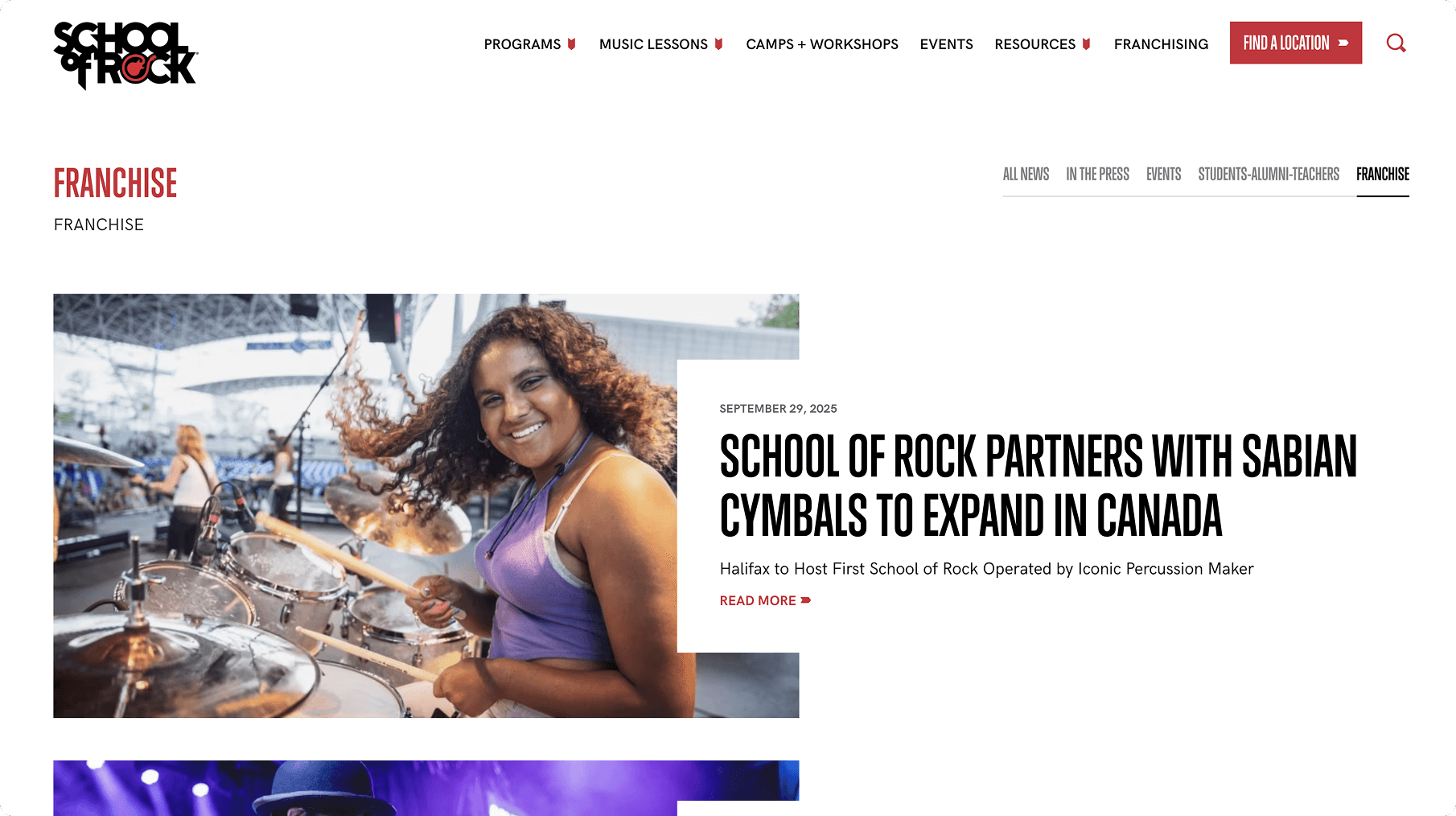 Smiling person with curly hair plays a drum kit on an outdoor stage, wearing a purple tank top.