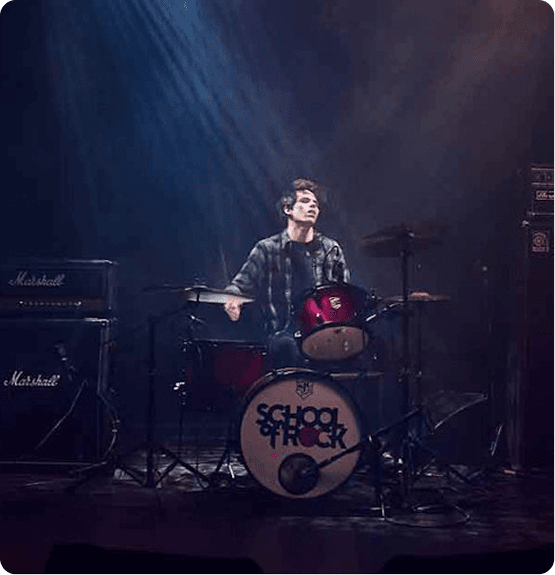 Drummer at a red drum kit on a dark stage with blue lighting; bass drum reads "School of Rock" while Marshall amps stand to the side.