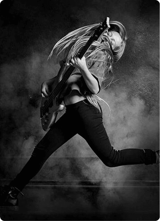 Black-and-white image of a musician mid-air playing a bass guitar, long hair flying, wearing dark clothes amid smoky stage lighting.