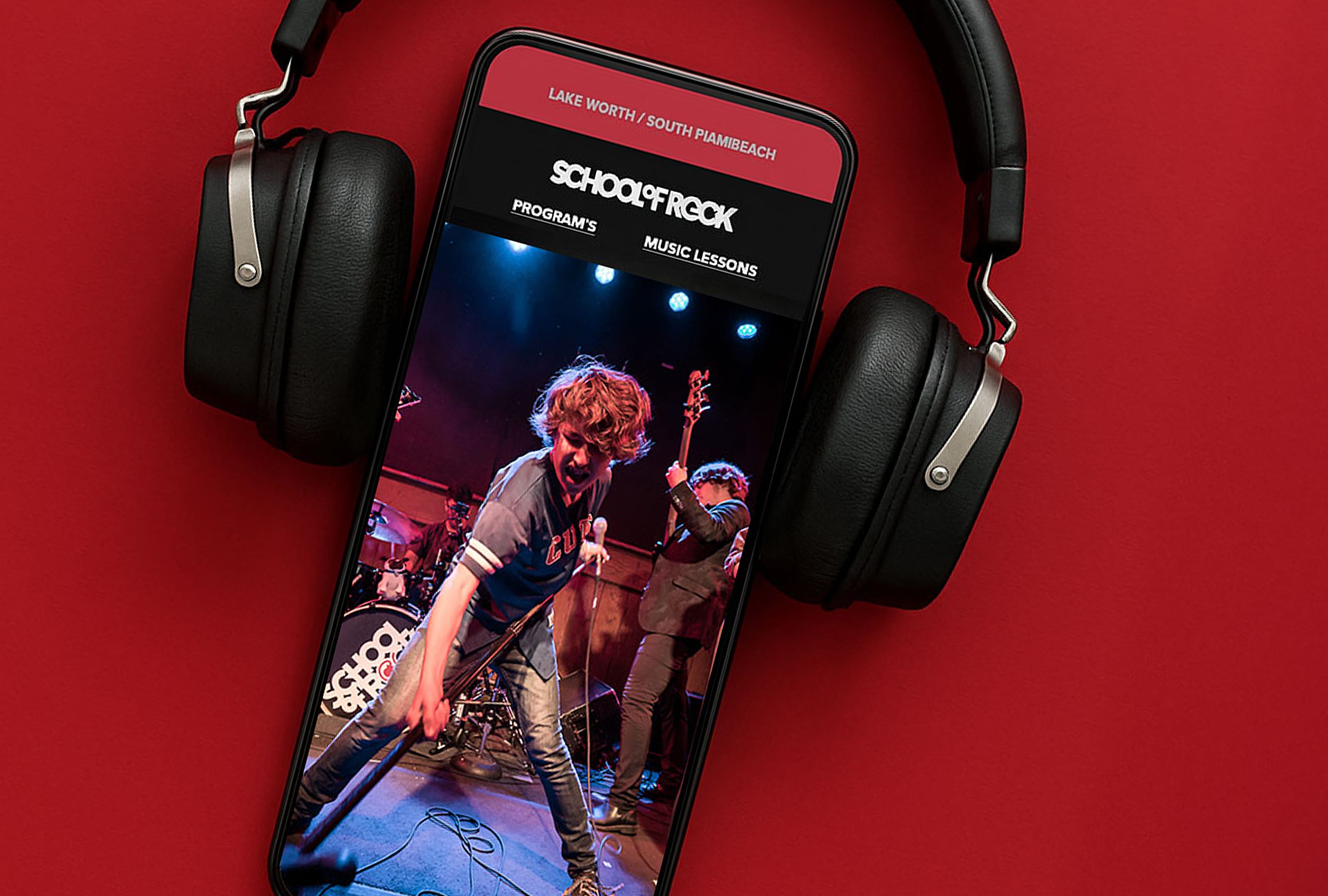 Smartphone on a red backdrop with over-ear headphones, displaying a live rock band performance and a guitarist mid-motion on stage.