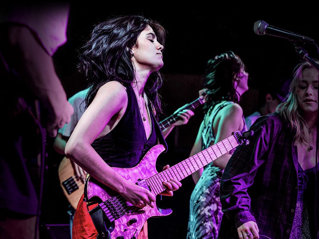 Guitarist with long dark hair in a sleeveless black top plays a purple electric guitar on a purple-lit stage, with other band members nearby.