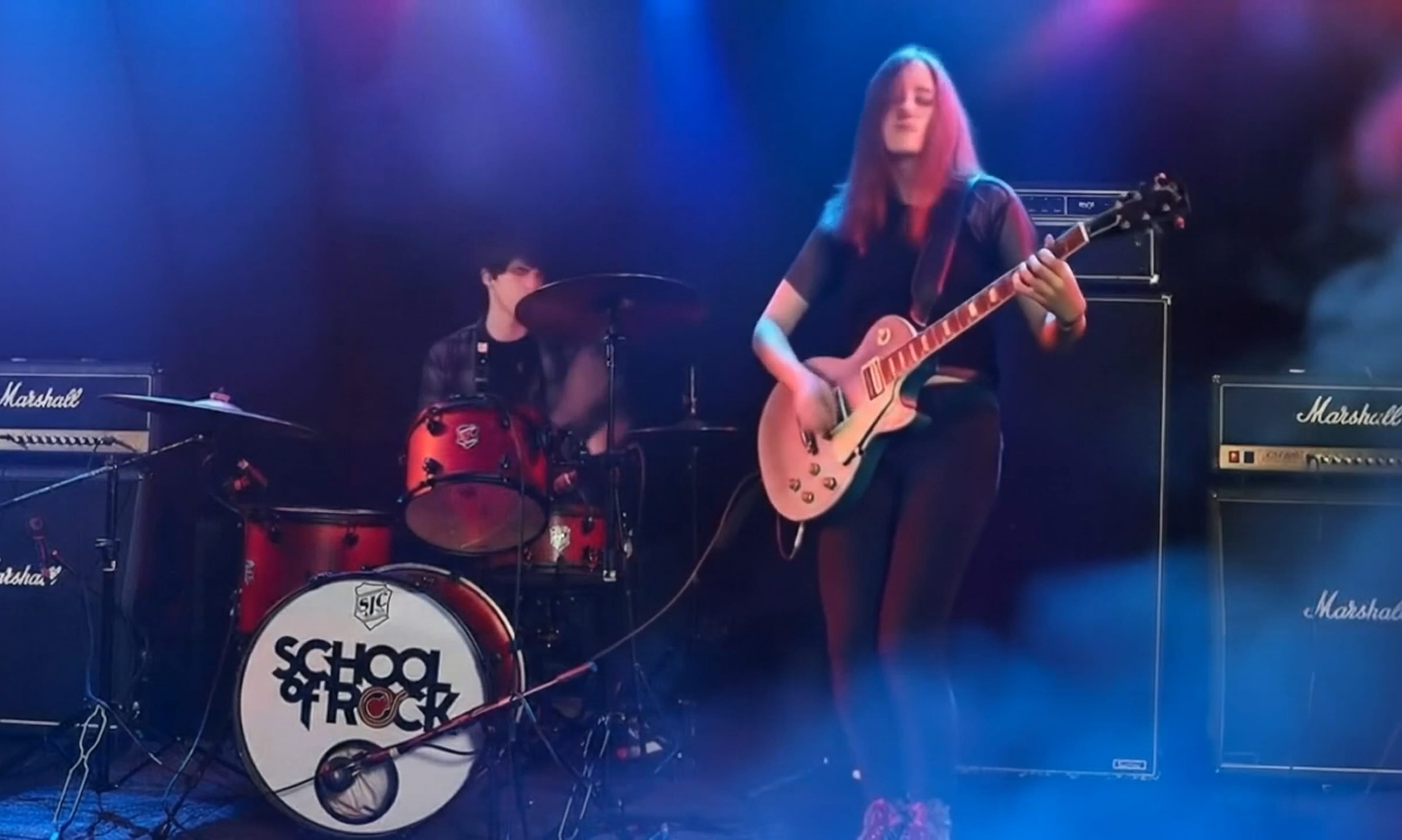 Long-haired guitarist in dark clothing plays a pale electric guitar on a smoky stage, with a red School of Rock drum kit behind and Marshall amps.