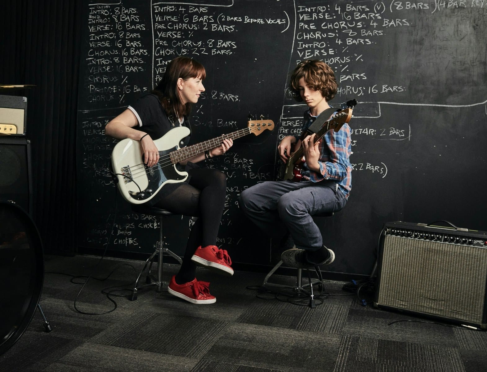 Two people sit on stools in a music room, playing bass and electric guitar, in front of a chalkboard wall filled with chords and verses.