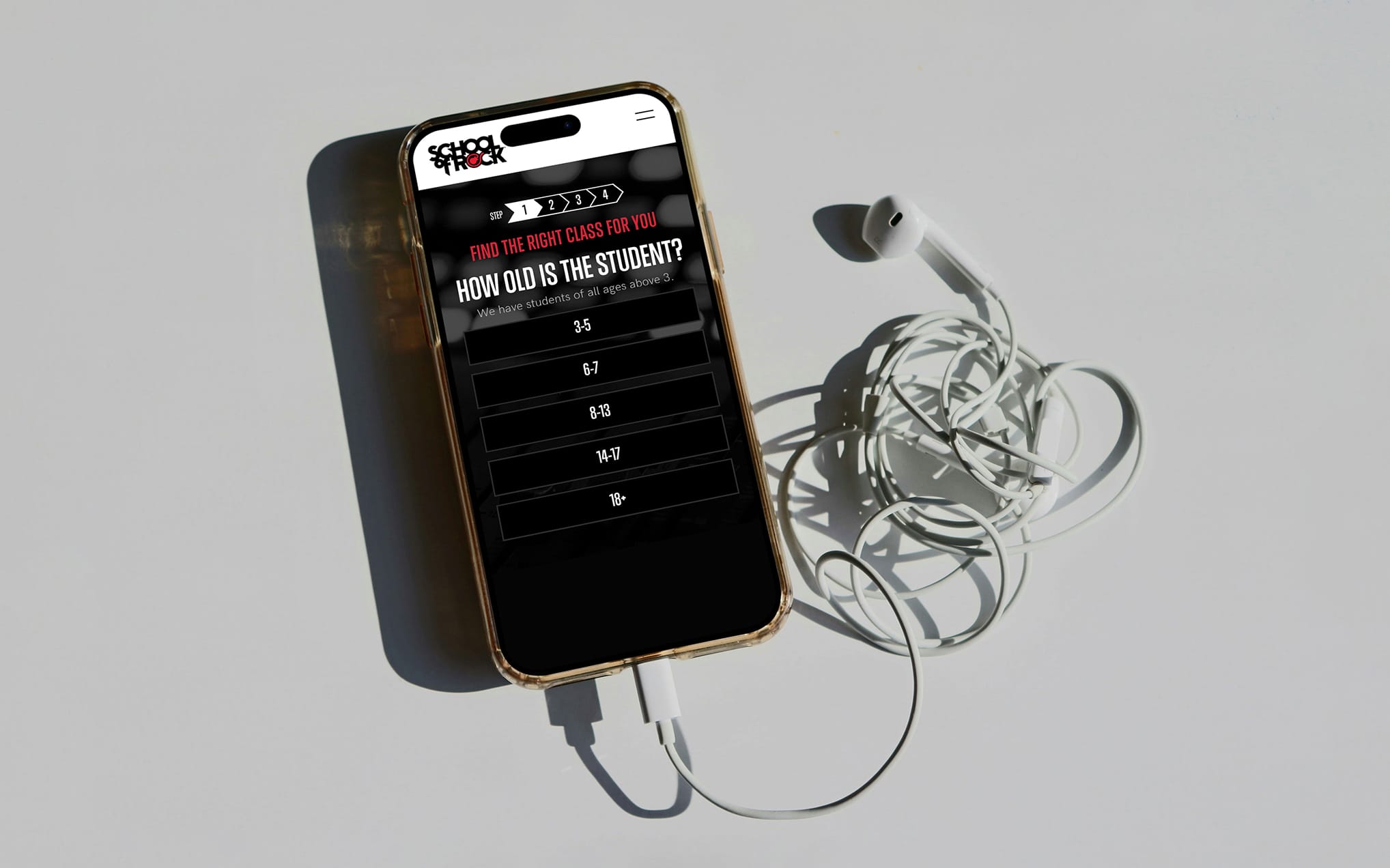 Phone in a brown case displays a quiz screen ('How old is the student?') with age options; tangled white earphones lie on a pale surface.