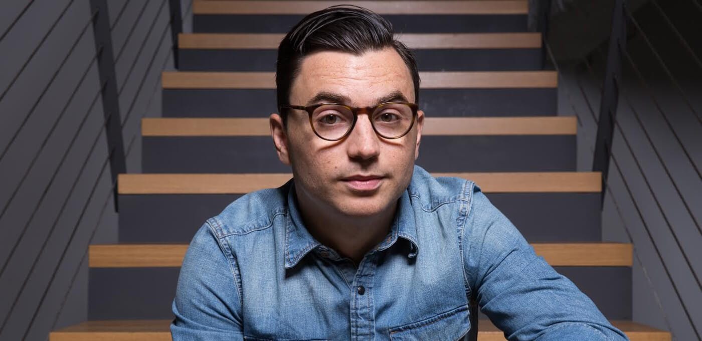 Person with short dark hair and round glasses wearing a denim shirt sits on wooden stairs, looking at the camera.