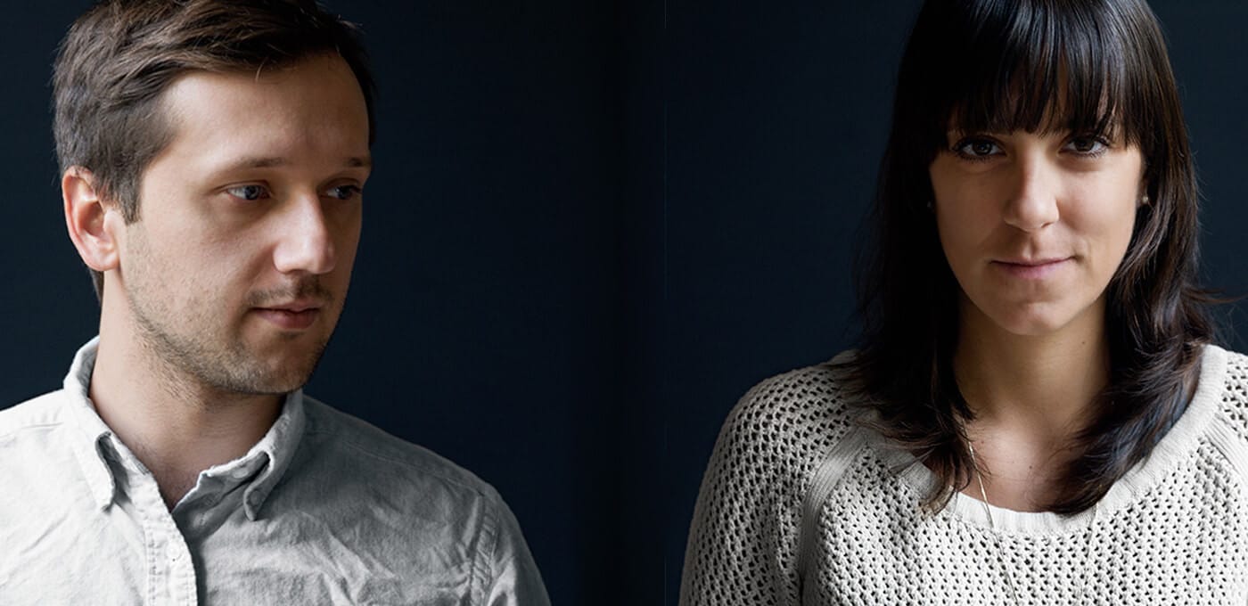Two people against a dark backdrop; left person in a pale button-down shirt looks toward the right, who wears a cream knit sweater and faces the camera.