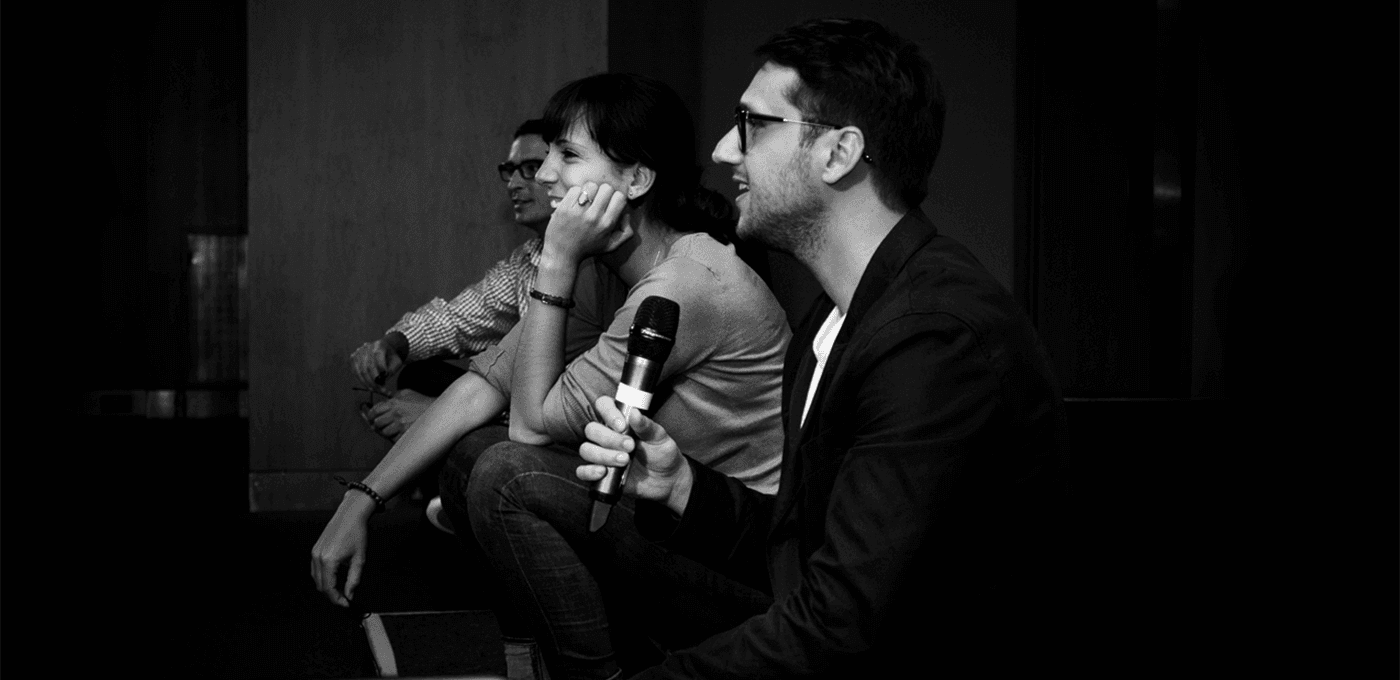 Black-and-white photo of four people seated in a row on a stage; the person on the right holds a microphone and smiles, while others listen.