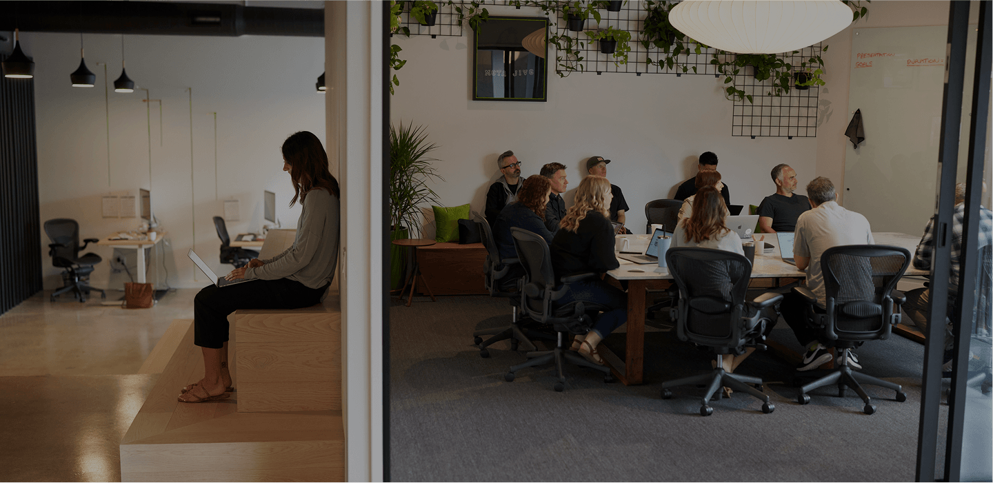 A person sits on wooden steps at the edge of open office, typing on a laptop; to the right, a large meeting room with a long table and many colleagues