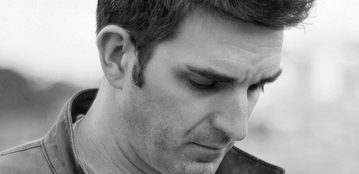 Close-up of a person with short dark hair looking downward, wearing a collared jacket; black-and-white portrait conveying a contemplative mood.