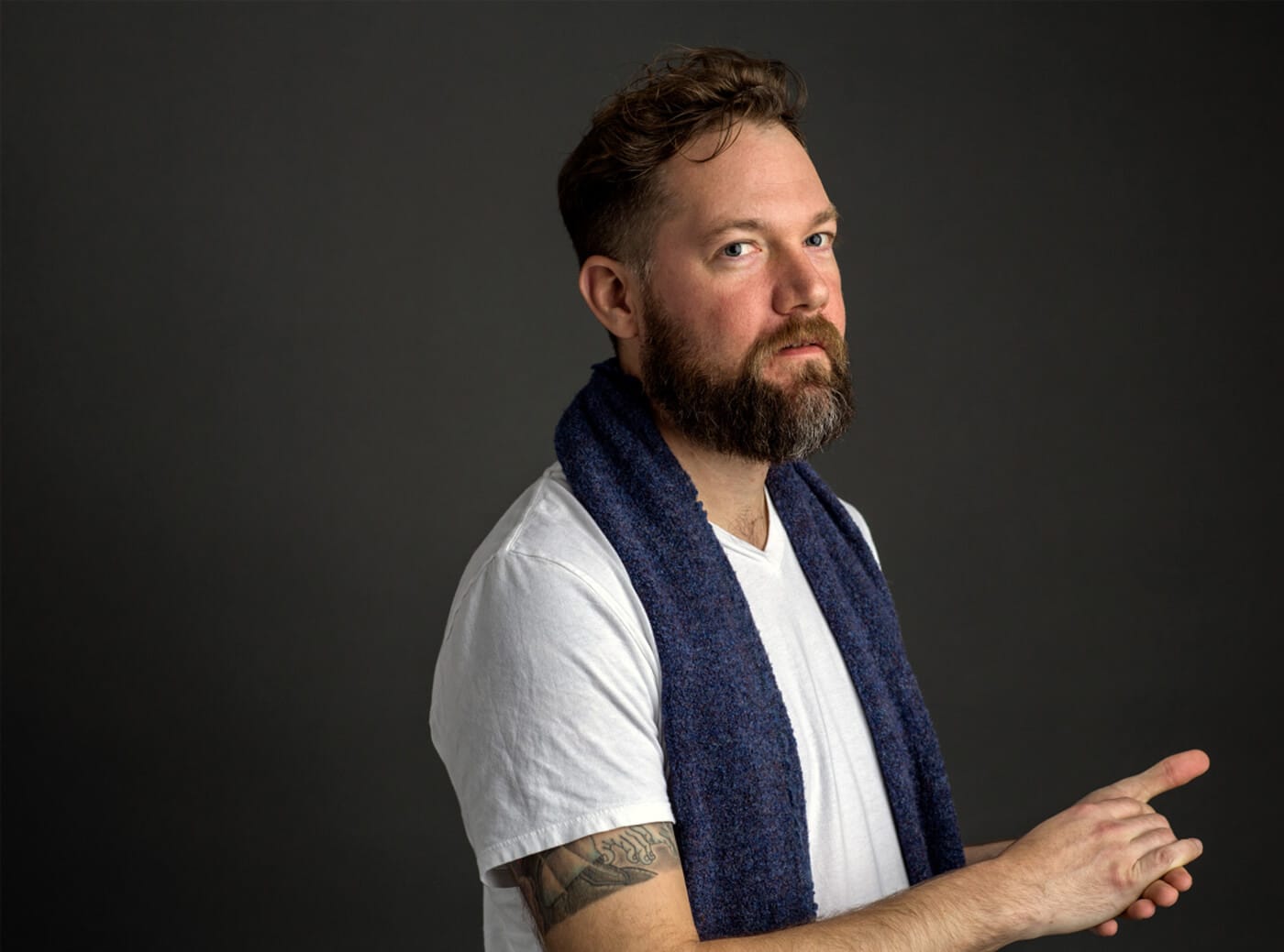Bearded person in a white T‑shirt with a dark blue scarf, against a dark backdrop; visible forearm tattoo and hands gesturing with an extended finger.