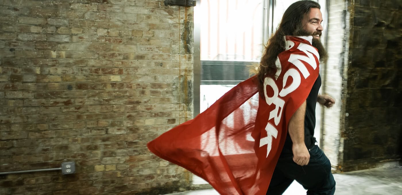 Person with long hair and a beard wears a red cape with bold white lettering, cape fluttering behind as they walk past a brick-walled industrial space.