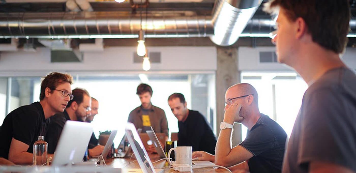 Group of people around a long conference table in a bright office, working on laptops; exposed ceilings and hanging lights, with a person on the right.