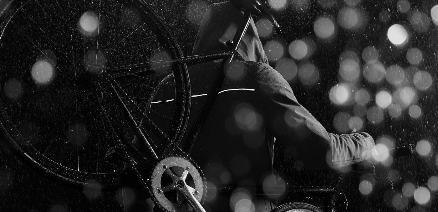 Person in a rain jacket carries a bicycle over their shoulder at night, with bright city lights and raindrop bokeh in grayscale.