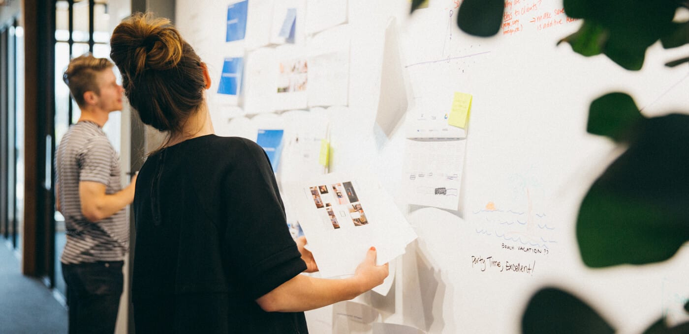 Person with a bun and black top examines papers on a wall covered with notes and sketches; another person in a striped shirt stands in the background.