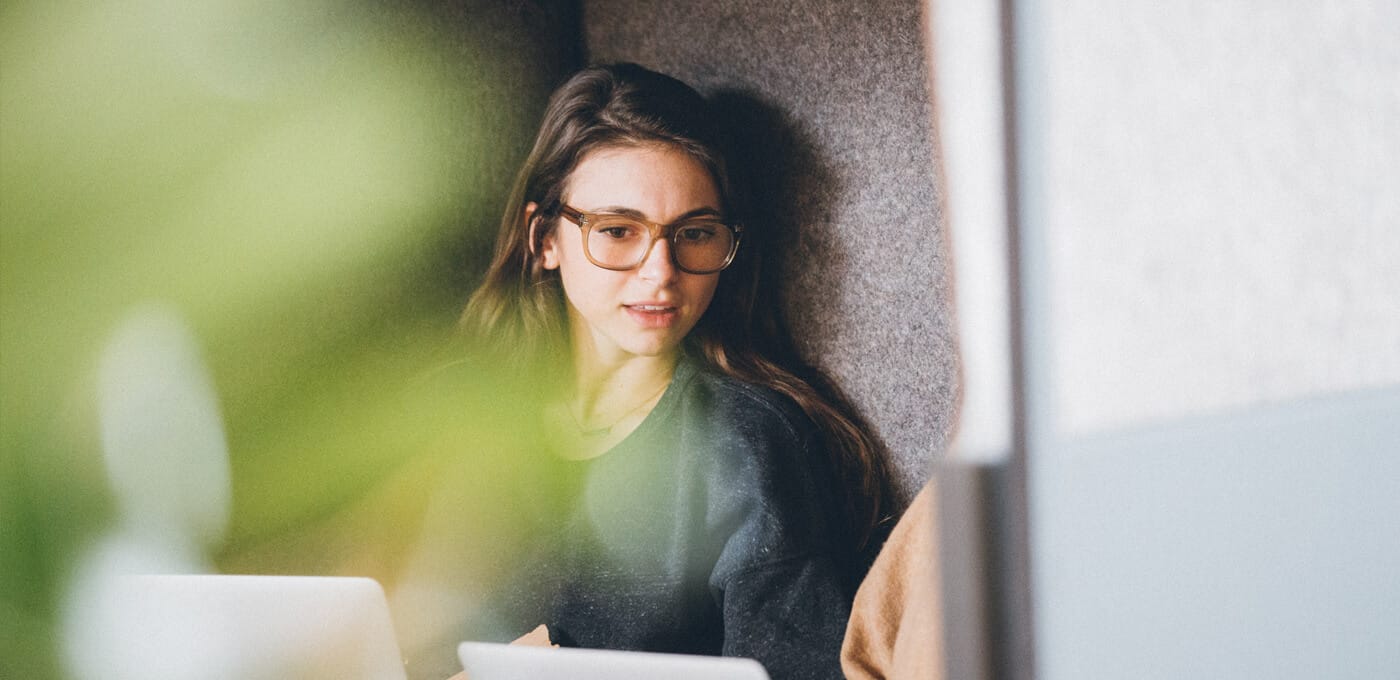 Person with long hair and brown glasses sits in a cozy corner, looking at a laptop; a blurred green plant in the foreground.