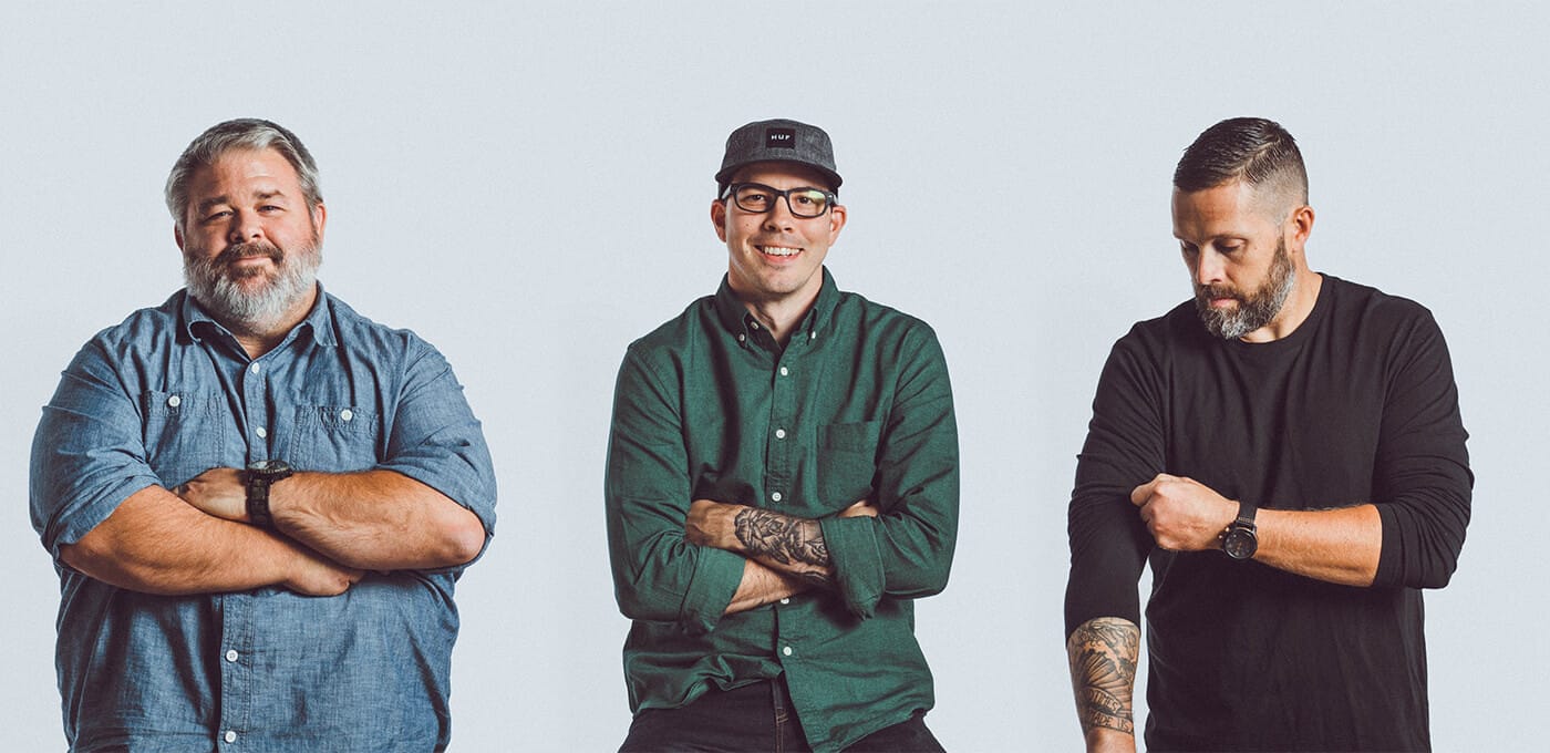 Three individuals stand with arms crossed against a plain light backdrop: left in a denim shirt, middle in green with cap and glasses, right in black with tattoos.