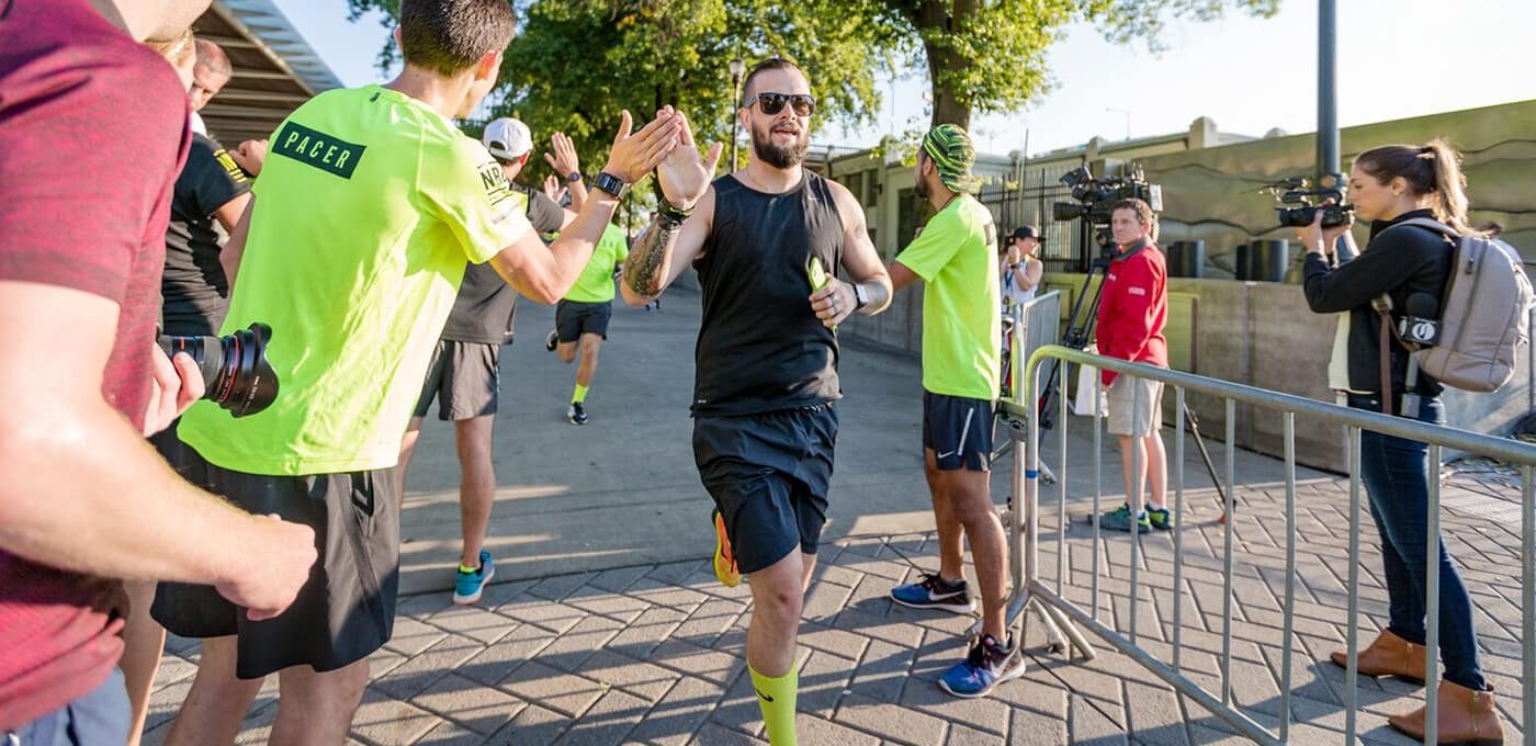 Bearded runner in a black sleeveless shirt high-fives volunteers in neon shirts as photographers and media capture the moment beside metal barriers.