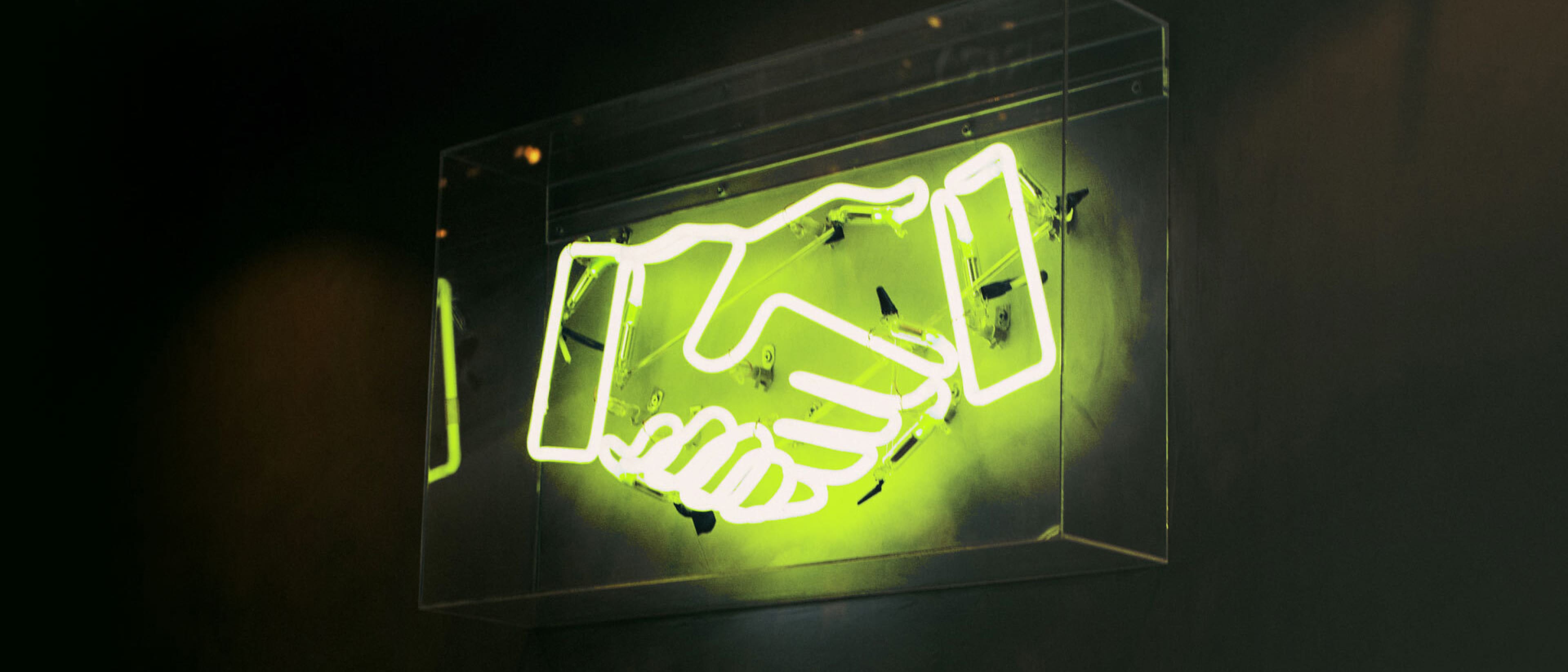Neon lime-green handshake sign enclosed in a clear display case mounted on a dark wall, glowing softly.
