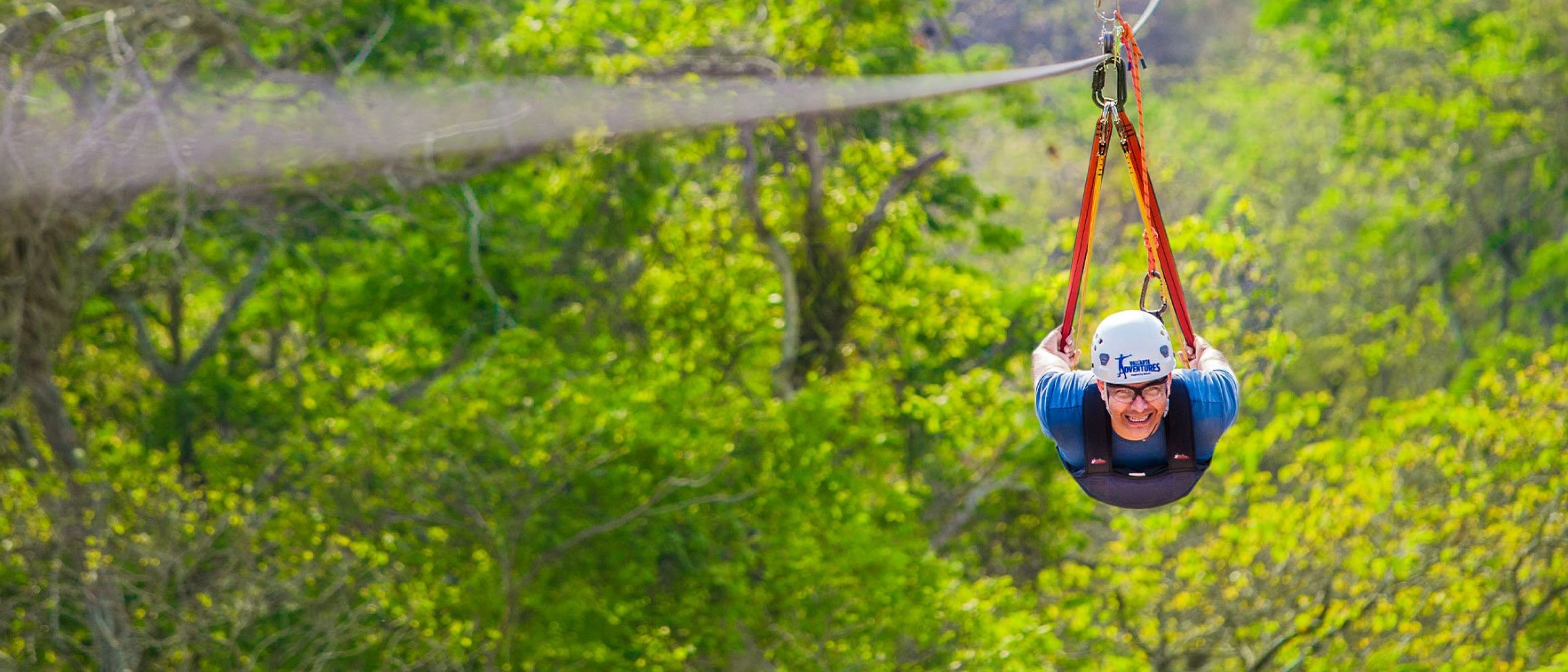 Person zip-lining through a lush green forest, wearing a white helmet and blue shirt, smiling as they glide along a harnessed zip-line.
