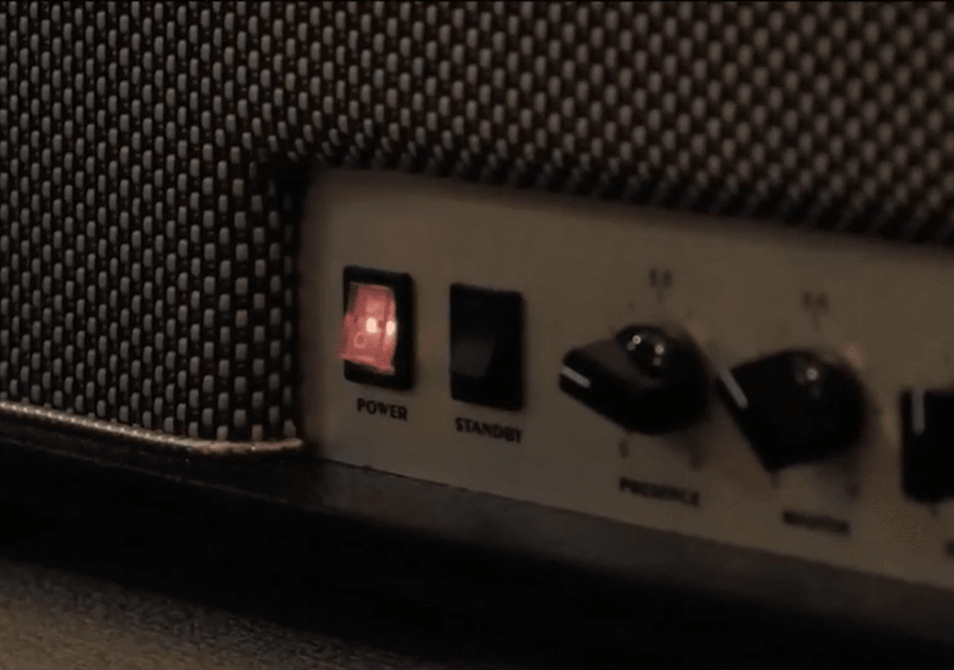 Close-up of a speaker front panel with a glowing red power switch, a standby switch, and two control knobs beside a black mesh grille.