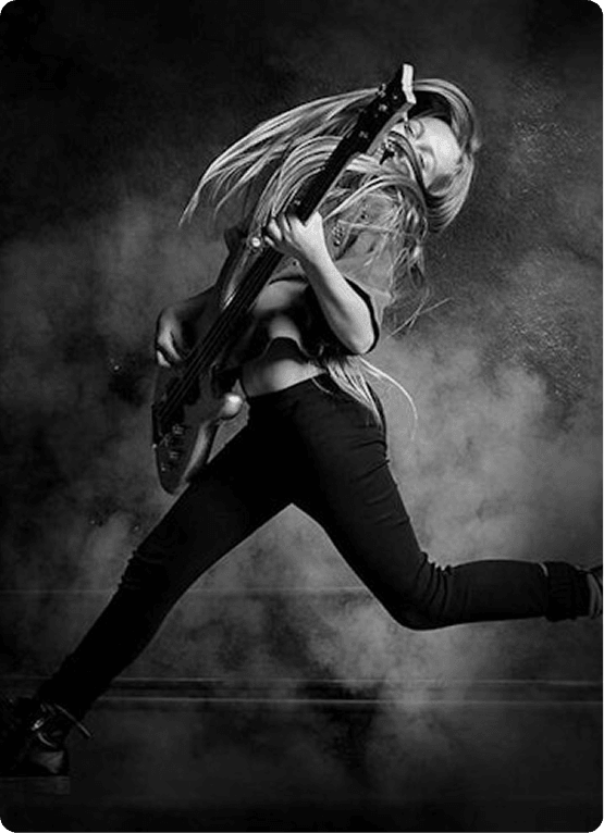 Black-and-white photo of a musician mid-air, energetically playing a bass guitar as long hair sweeps across their face, on a smoky stage.