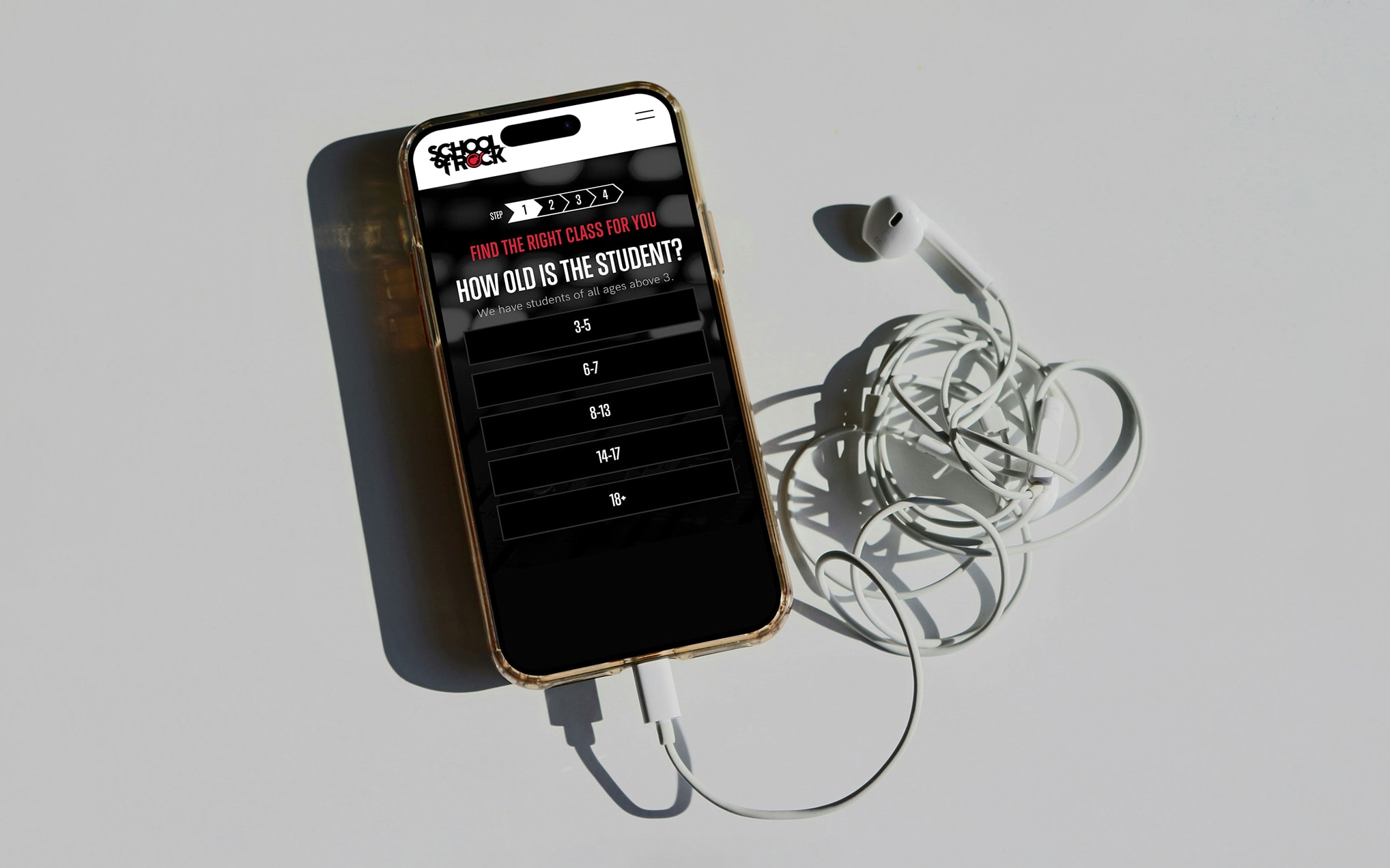 Gold smartphone displays a quiz screen reading 'HOW OLD IS THE STUDENT?' with age options, beside tangled white earphones on a light gray surface.