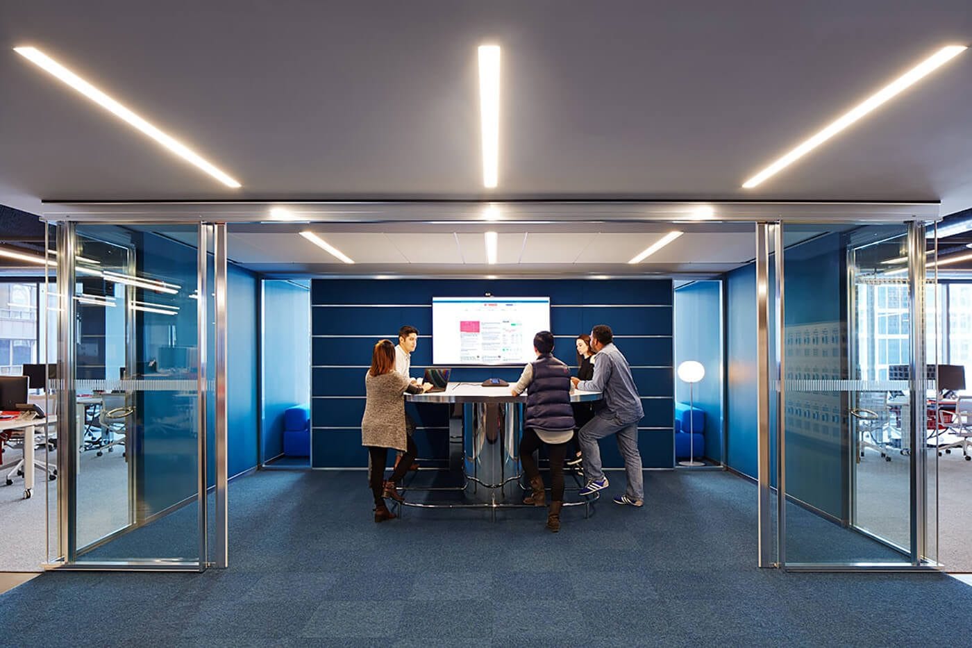 Five people stand around a high table in a modern glass-walled meeting room, looking at a wall screen displaying a presentation against blue panel walls.