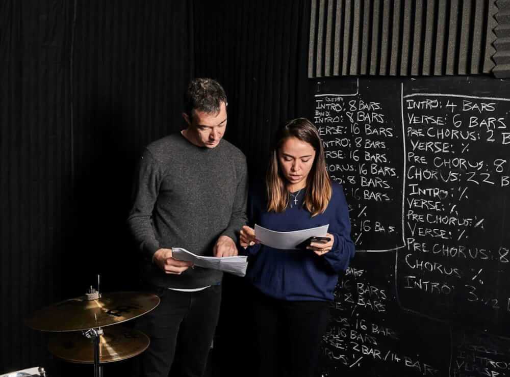 Two people stand in a rehearsal room with black walls and a chalkboard covered in musical notes and bars, reading papers beside a drum set.