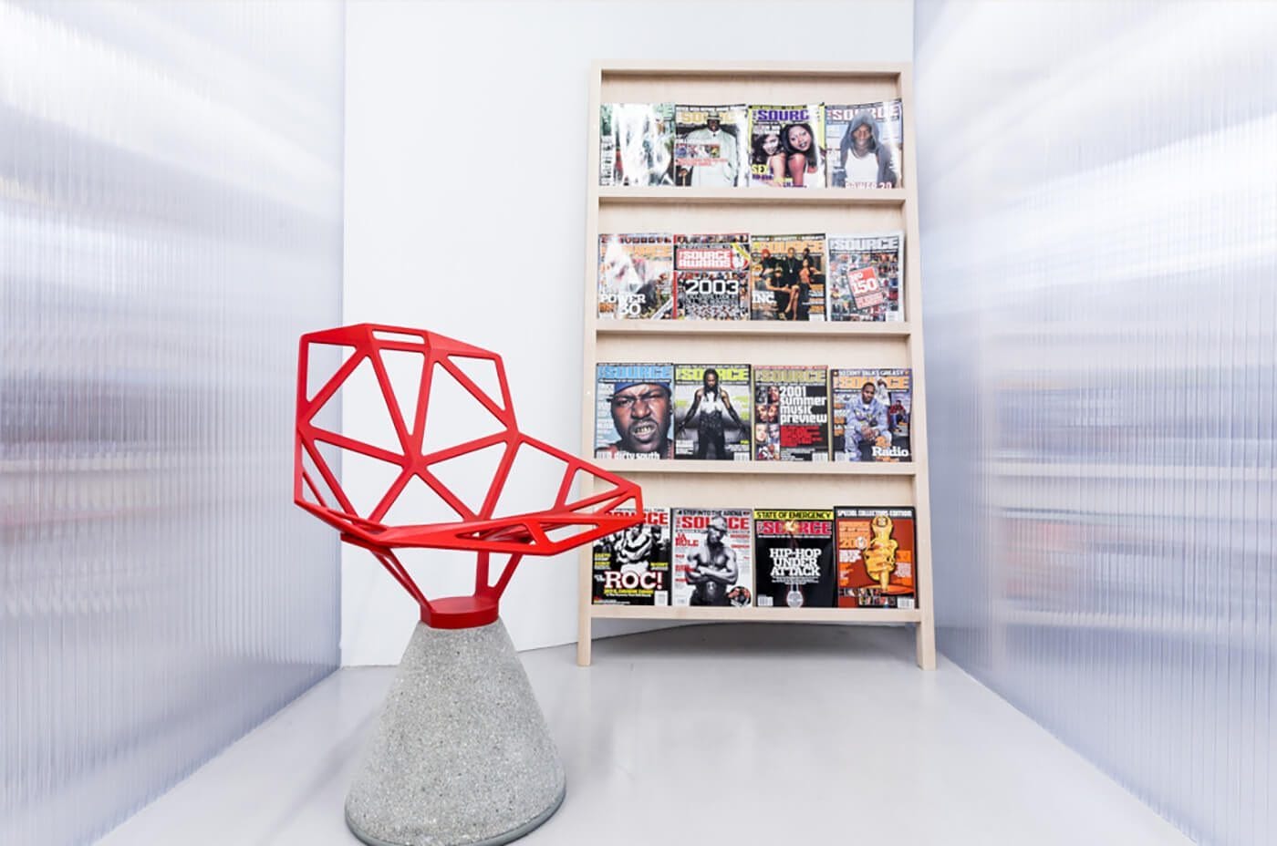 Modern room with a geometric red chair on a concrete cone pedestal beside a tall wooden magazine rack filled with colorful magazine covers.