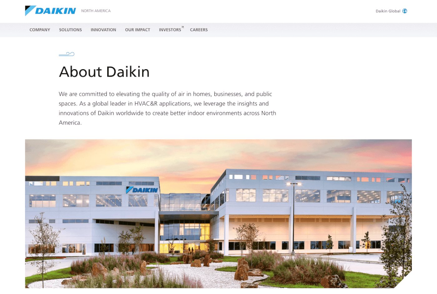 Daikin corporate building with a modern white facade and glass windows, a rock garden and landscaped foreground, Daikin logo on the left, under a warm sunset sky.
