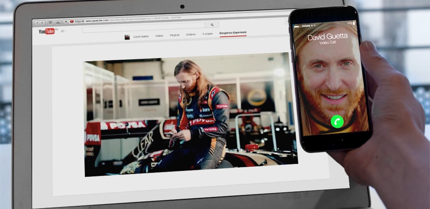 Laptop screen shows YouTube with a racer in a garage; a hand holds a smartphone displaying a video call with a smiling bearded man.