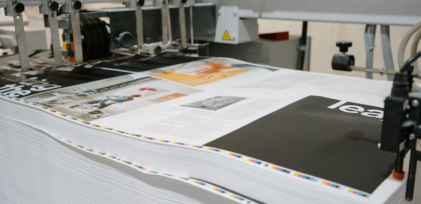 Close-up of a printing press feeding large sheets with images and text; edges show color registration marks, with machinery and rollers in view.