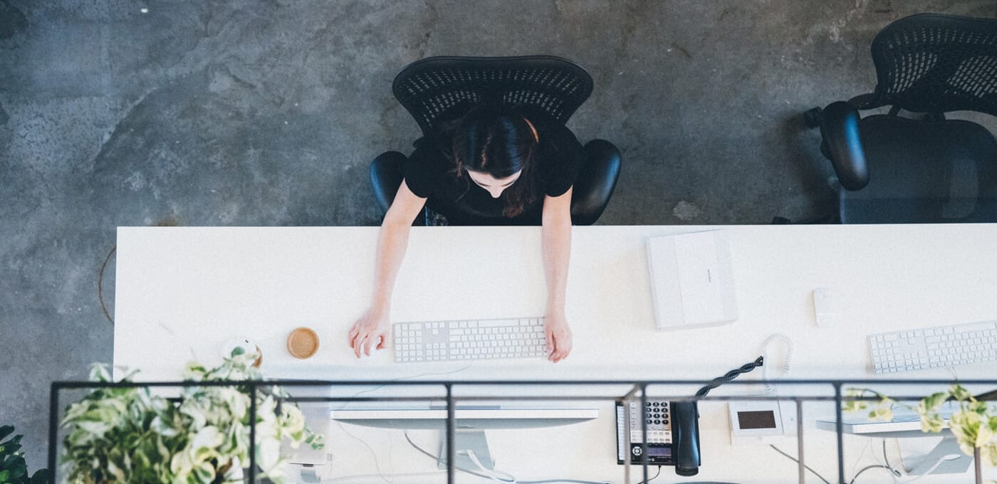 Top-down view of a person in a black top seated at a white desk, typing on a keyboard; coffee cup, notebook, phone, and plants on the desk.