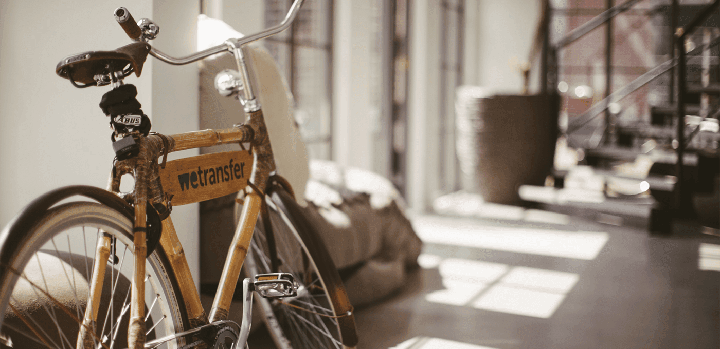 Wooden bicycle with a nettransfer sign on the frame sits indoors beside a couch and a large planter, bathed in sunlight with striped floor shadows.