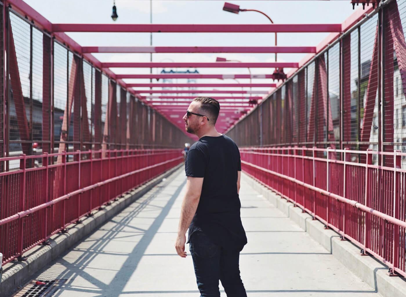 A person wearing a black shirt and dark jeans walks along a pink pedestrian bridge with red railings and mesh sides, wearing sunglasses.