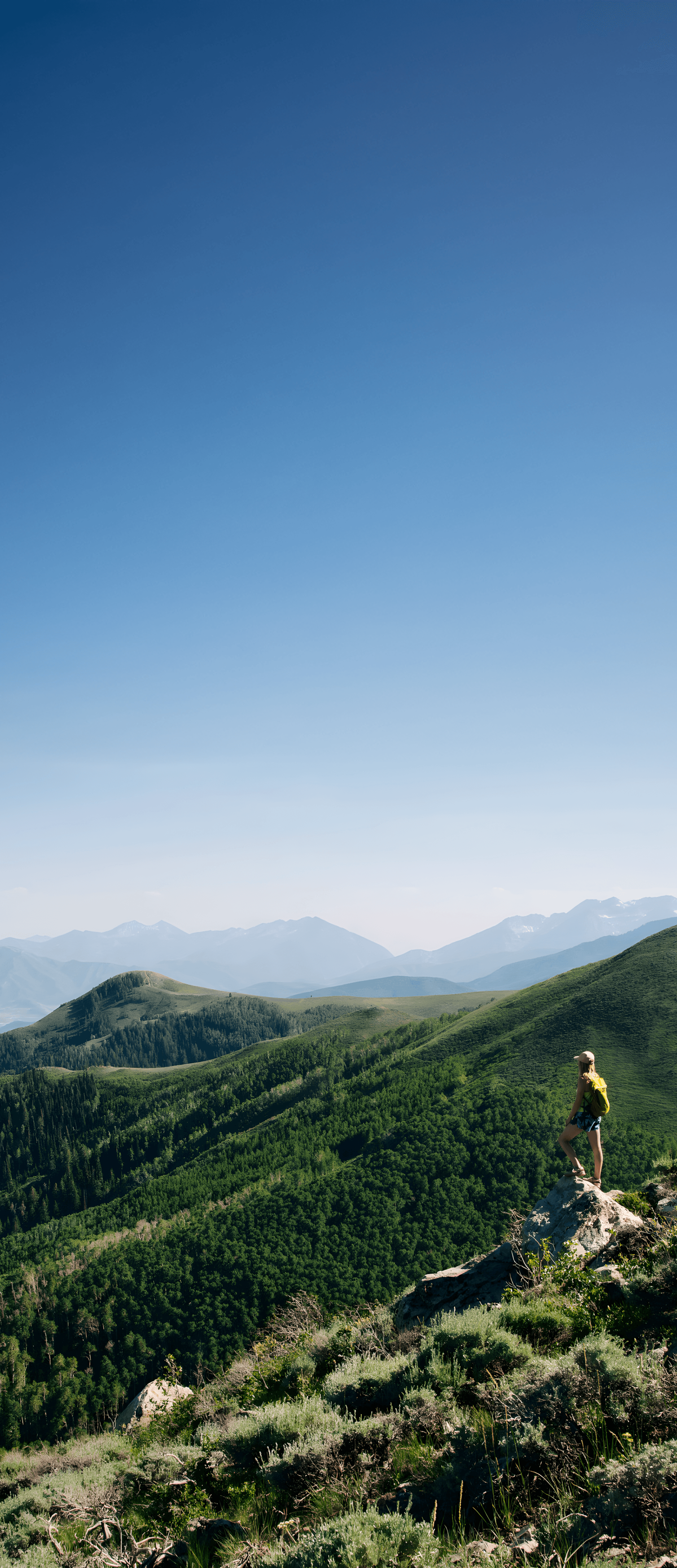 Hiker with a yellow backpack stands on a rocky outcrop, overlooking green rolling hills and distant mountains under a clear blue sky.