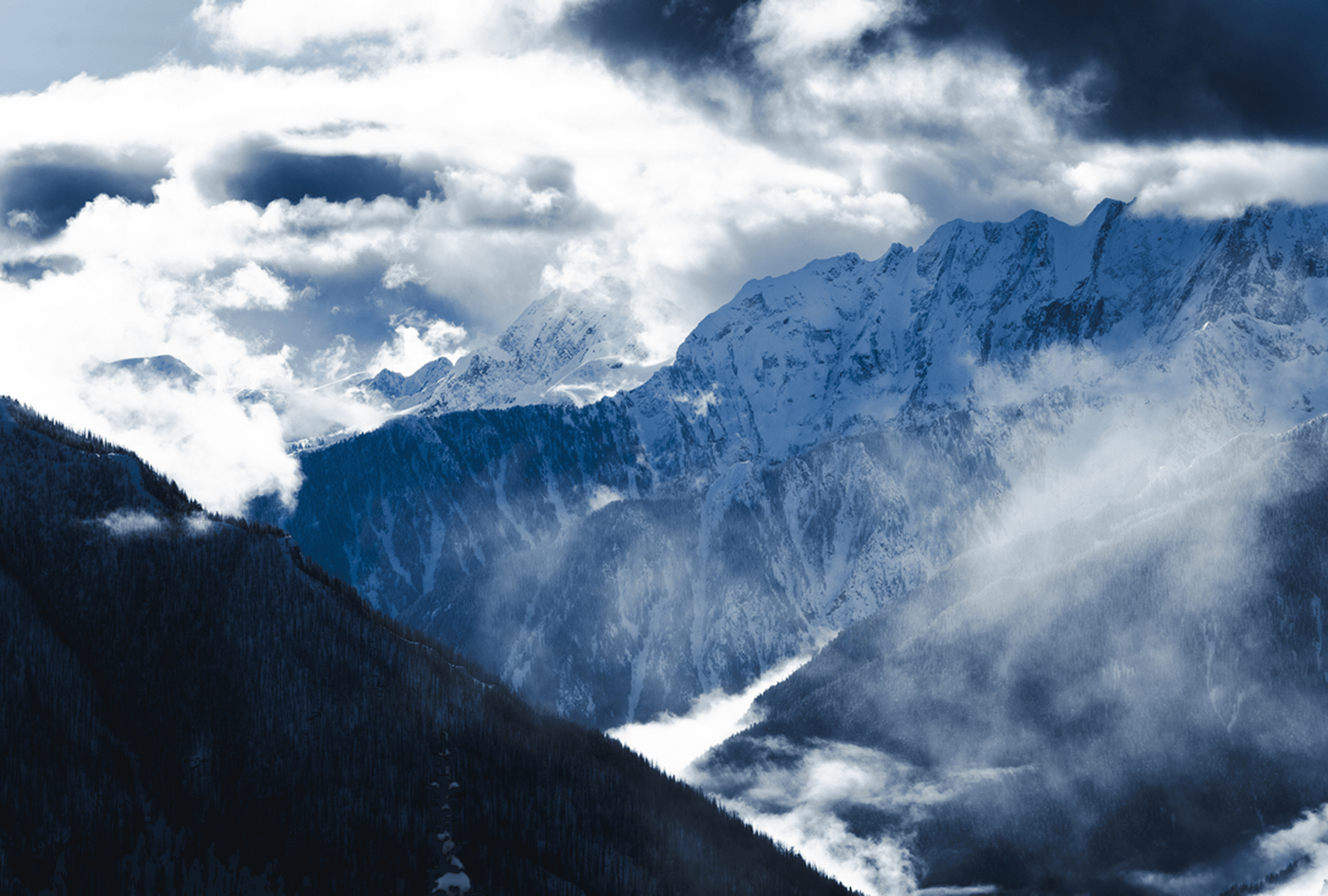 Snowy alpine mountains under dark blue clouds; mist fills the valley as forested slopes descend.