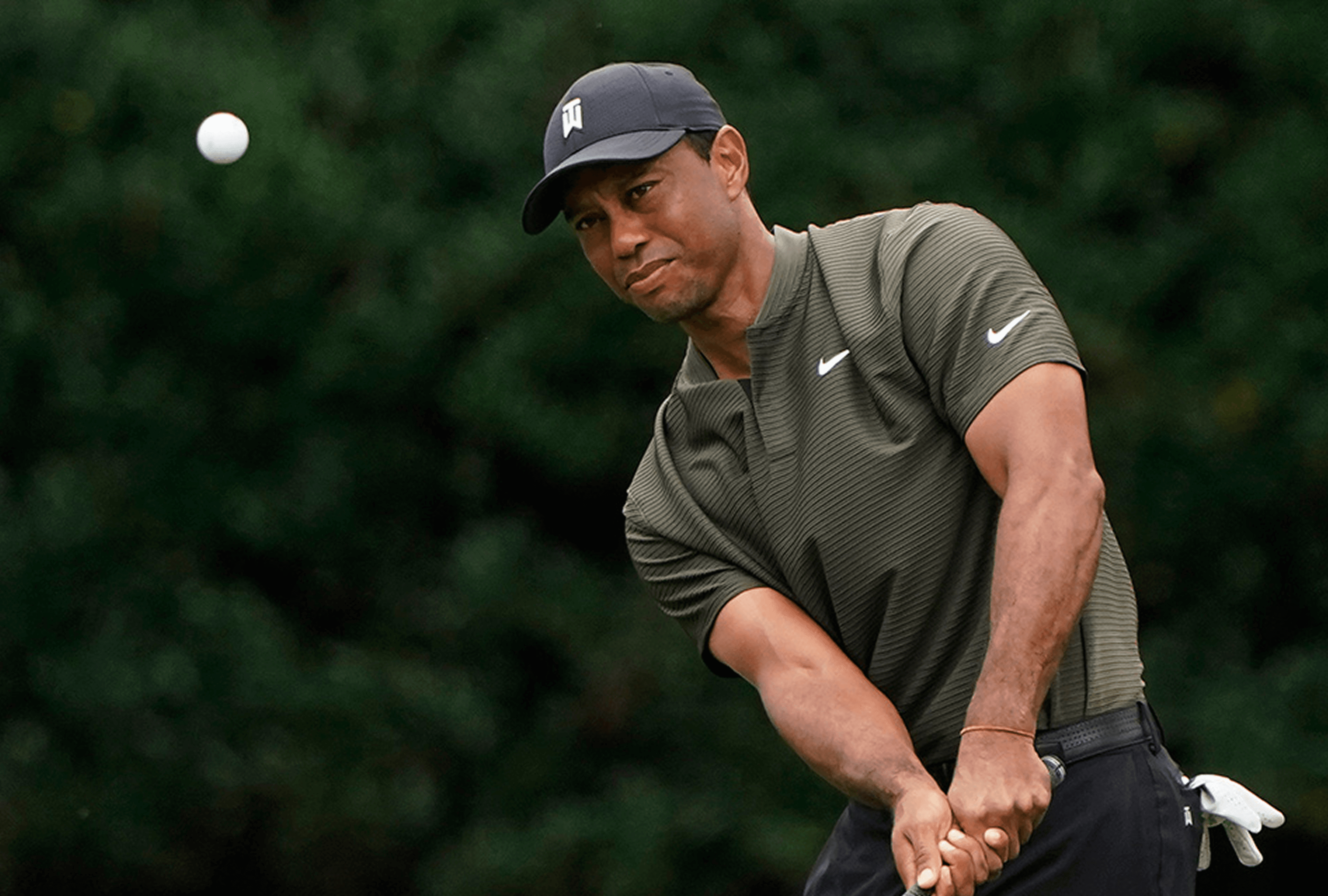 A golfer in a dark cap and olive polo swings, watching the white ball fly against a leafy green backdrop.