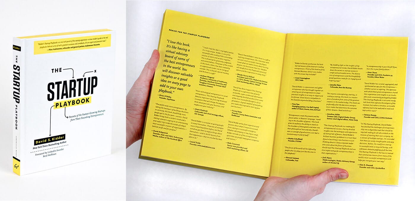 White hardcover of The Startup Playbook stands upright beside an open bright yellow book with dense black text; a hand holds the yellow pages.