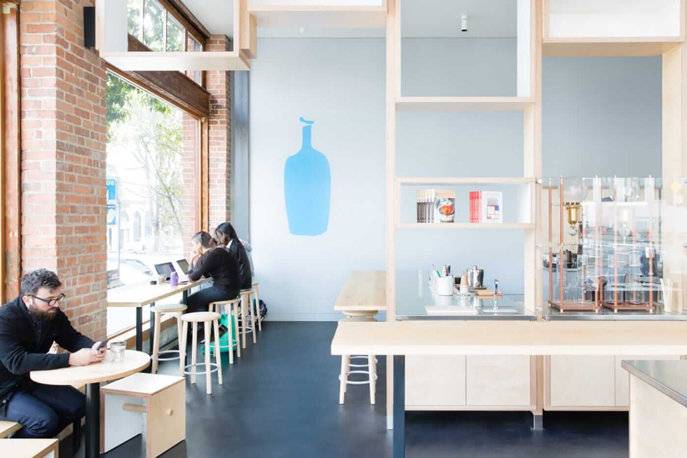 Bright cafe with brick wall and windows; a person with glasses sits at a round table using a phone, blue bottle mural on pale wall.