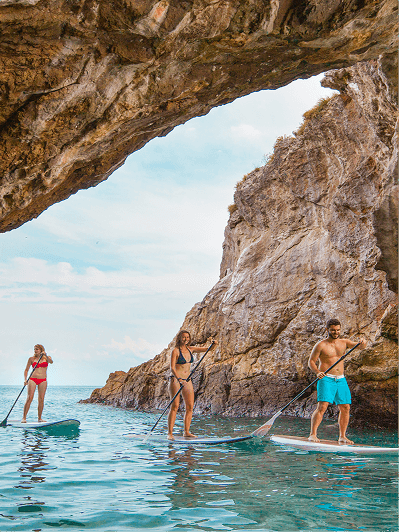 Three people stand-up paddleboarding beneath a rocky coastal arch, turquoise water and a blue sky.