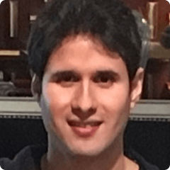Portrait of a person with short dark hair, smiling at the camera, wearing a dark shirt, indoors with a blurred background.