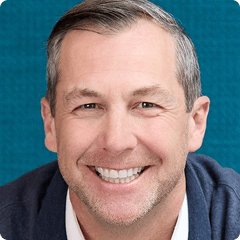 Smiling person with short gray hair, wearing a dark jacket over a white shirt, against a teal textured background.
