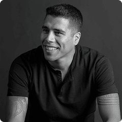 Grayscale portrait of a smiling person in a dark short-sleeve shirt; visible forearm tattoos, facing slightly left against a neutral backdrop.