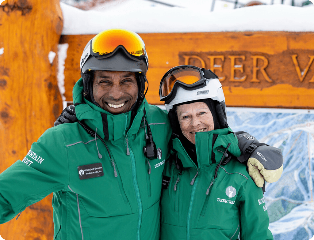 Two skiers in green jackets hug and grin in front of a wooden Deer Valley sign on a snowy day, wearing helmets and goggles.