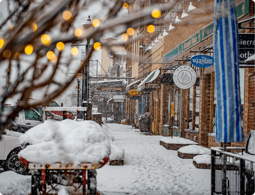 Snowy street with brick shops, signs, and a blue flag; blurred string lights in the foreground, snow-covered tables, and a parked van.